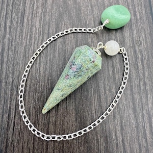 Peut inclure: Un pendule en pierre précieuse verte et rouge avec une chaîne argentée. Le pendule a un cristal en forme de cône facetté, une petite sphère blanche et une pierre ovale verte. La chaîne est argentée et relie les pierres.