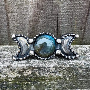 May include: A silver-toned barrette featuring a central, round, iridescent blue-green stone, flanked by crescent moon shapes. The barrette is adorned with small, round silver accents and a beaded border, creating a celestial-themed accessory.