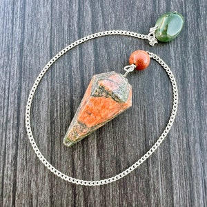 Puede incluir: Un péndulo de cristal rojo, verde y blanco con una cadena plateada y una piedra verde en la parte superior.