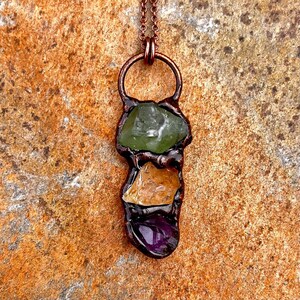 Puede incluir: Un collar con un colgante de alambre de cobre envuelto alrededor de tres piedras preciosas de diferentes colores. La piedra superior es verde, la piedra del medio es naranja y la piedra inferior es morada.