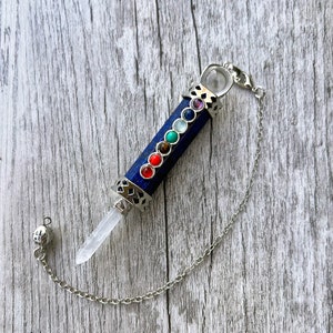 May include: A silver and blue crystal pendulum with a clear quartz point and a chain. The pendulum has seven colored stones representing the seven chakras.
