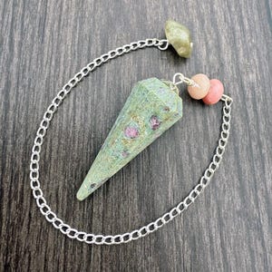 May include: A green and pink gemstone pendulum with a silver chain. The pendulum has a faceted, cone-like shape with pink inclusions. The chain connects to a small, light green stone and two pink beads.