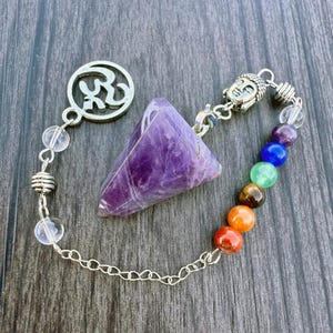 May include: Amethyst crystal pendulum with a silver chain and a silver charm with the Om symbol. The chain also has clear quartz beads and seven chakra beads in the colors red, orange, yellow, green, blue, indigo, and violet.