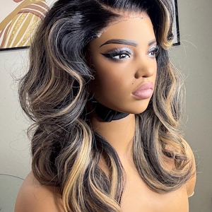 May include: A mannequin head displaying a long, wavy wig with a side part. The wig features a blend of black, brown, and blonde highlights. The mannequin has makeup, including silver eyeshadow and dark eyeliner. A black choker is around the neck.