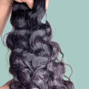 May include: A bundle of dark, wavy hair extensions held against a light teal background. The hair appears to be a deep black with subtle gray undertones, showcasing a natural wave pattern. The texture is soft and flowing.