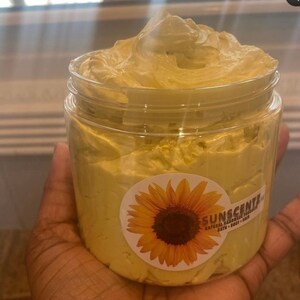May include: A clear jar filled with a yellow, creamy body butter. The jar has a label with a sunflower and the text "SUNSCENTS NATURAL HAIR & BODY PRODUCTS BATH - BODY - HAIR".