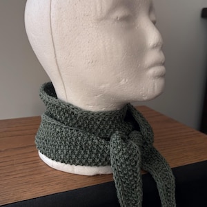 May include: A dark olive green crocheted neck scarf featuring a textured, dense stitch pattern. The scarf is tied in a simple knot at the front, with two tapered ends hanging down. The accessory is displayed on a white, featureless mannequin head.