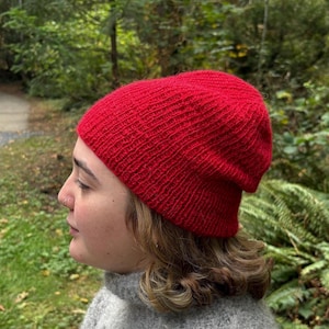 May include: A vibrant red knit beanie hat featuring a classic ribbed texture. The hat is constructed with a folded brim and a slightly slouchy fit, showcasing a consistent vertical stitch pattern throughout the entire surface of the headwear.