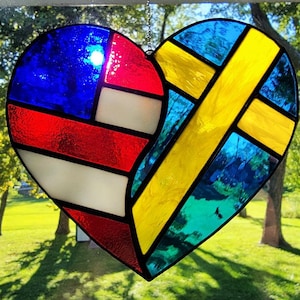 Puede incluir: Un corazón de vidriera que combina las banderas estadounidense y sueca. El lado izquierdo presenta secciones rojas, blancas y azules, mientras que el lado derecho muestra una cruz amarilla sobre un fondo azul. El corazón está suspendido, con un fondo verde y soleado.