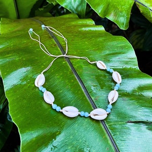 Puede incluir: Un collar hecho a mano con conchas de cauri blancas y cuentas azul claro, ensartadas en un cordón blanco. El collar se muestra sobre una gran hoja verde brillante, creando una estética natural y playera.