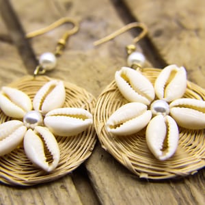 Könnte beinhalten: Ein Paar Ohrringe aus geflochtenem Stroh und weißen Kaurischnecken. Jeder Ohrring hat ein blumenförmiges Design mit einer Perle in der Mitte.