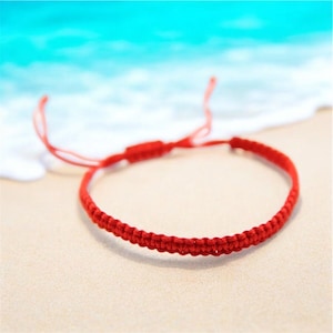 Puede incluir: Un brazalete de cuerda roja con un diseño de nudo simple. El brazalete está en una playa de arena blanca con agua azul de fondo.