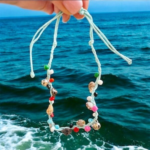 May include: A light blue braided bracelet with colourful beads and small seashells. The bracelet is held against a blue ocean background.