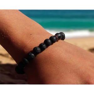 Könnte beinhalten: Ein Armband aus schwarzen Lavasteinperlen. Das Armband besteht aus runden, porösen schwarzen Perlen. Das Armband wird an einem Handgelenk getragen, mit einem Strand und dem Ozean im Hintergrund.