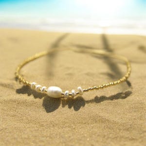 Op de afbeelding: Een delicate gouden ketting met kleine gouden kralen en een enkele witte parel hanger. De ketting ligt op een zandstrand.