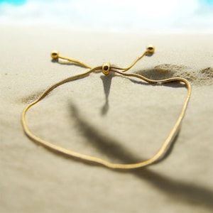 May include: A gold chain bracelet with a sliding knot clasp. The bracelet is lying on a sandy beach.