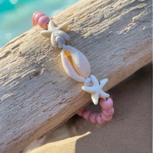 以下が含まれることがあります： ピンクと白のビーズブレスレットに、白い貝殻とヒトデのチャームが付いています。ブレスレットは流木の上に置かれています。