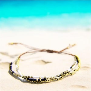 May include: A layered bracelet with white, black, and gold beads on a brown cord. The bracelet is on a white sandy beach.