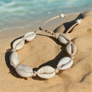 May include: A white seashell bracelet with a natural rope cord. The bracelet is made of small, white seashells strung together with a natural rope cord. The bracelet is lying on a sandy beach.
