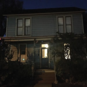 May include: A two-storey blue house with a porch and a lit front door. The house has white trim and windows with curtains. The house is surrounded by trees and bushes.