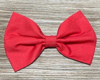 May include: A solid coral-red fabric bow tie featuring a classic butterfly shape with a central knot. The textile has a visible woven texture and a matte finish, with symmetrical folded wings extending outward from the center piece.