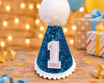 May include: A cone-shaped party hat covered in blue glitter with a large white pom-pom on top. The hat features a silver-colored number one patch centered on the front and is trimmed along the bottom edge with a white pom-pom fringe border.