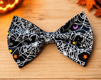 May include: A black bow tie featuring a white spider web pattern with small yellow, orange, and purple stars and crescent moons scattered across the fabric. The bow tie has a structured, symmetrical shape with a central knot and two pointed wings.