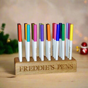 May include: A wooden pen holder with the name "FREDDIE'S PENS" engraved on the front. The holder has 20 slots for pens and is filled with colourful markers.