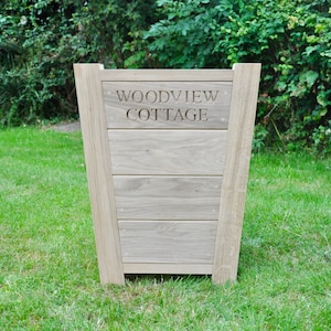 May include: A light-coloured wooden planter box with the engraved text "WOODVIEW COTTAGE." The planter has a rectangular shape with a slightly tapered design and is set on a grassy lawn. The wood has a natural grain.