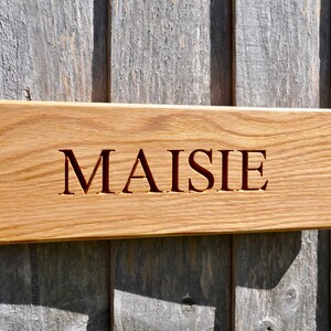May include: A wooden sign with the name "MAISIE" carved into it. The sign is made of light brown wood and has a natural finish.