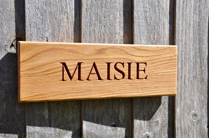 May include: A wooden sign with the name "MAISIE" carved into it. The sign is made of light brown wood and has a natural finish.