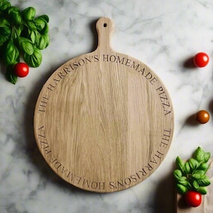 May include: A circular wooden pizza board with a handle, engraved with the text "THE HARRISON'S HOMEMADE PIZZA" around the edge. The board is light brown with a natural wood grain. Fresh basil and tomatoes are in the background.