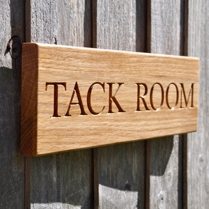 May include: A wooden sign with the words "TACK ROOM" carved into it. The sign is mounted on a wooden wall.