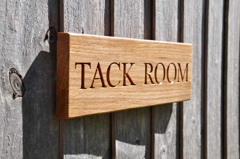 May include: A wooden sign with the words "TACK ROOM" carved into it. The sign is mounted on a wooden wall.