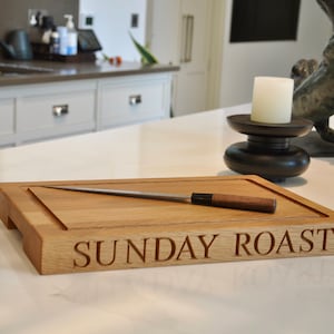May include: Oak cutting board with a carving knife, engraved with "SUNDAY ROAST". The board has a recessed area and a handle. A candle holder and sculpture are in the background. The board is likely 30 cm wide.