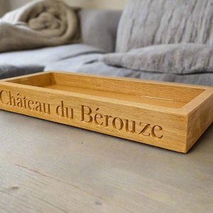 May include: A wooden tray with the words "Château du Bérouze" engraved on the front. The tray is made of light brown wood and has a rectangular shape.
