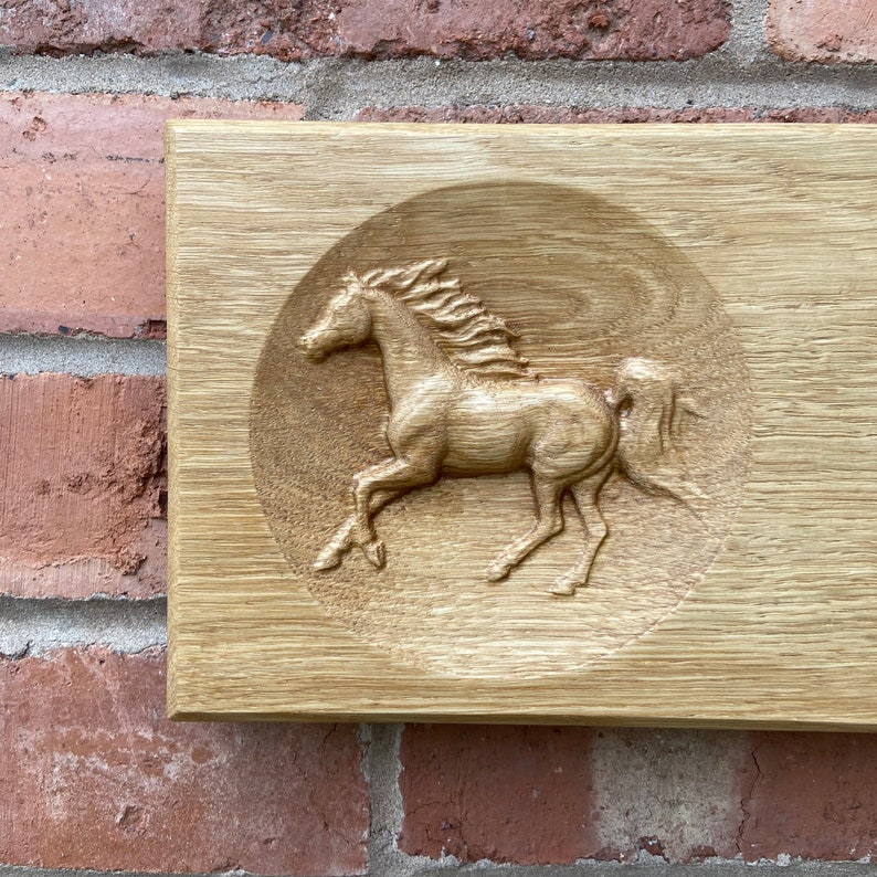 May include: A wooden plaque with a carved image of a horse running in a circle. The horse is carved in relief and has a light brown color.