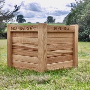 May include: A wooden box with the words "GRANDAD'S 60th BIRTHDAY" engraved on it. The box is made of light-coloured wood and is set on grass, with trees and a cloudy sky in the background. The box is approximately 60 cm in height.