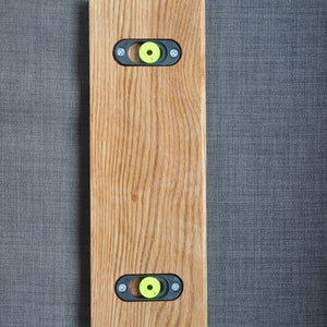 May include: A wooden wall mount with two black metal brackets and yellow plastic inserts. The mount is made of light brown wood with a natural grain pattern.
