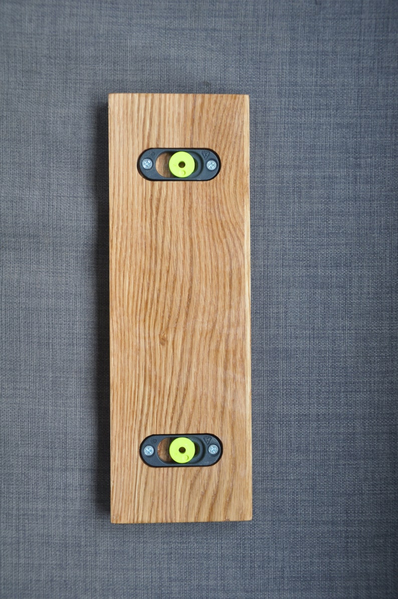 May include: A wooden wall mount with two black metal brackets and yellow plastic inserts. The mount is made of light brown wood with a natural grain pattern.