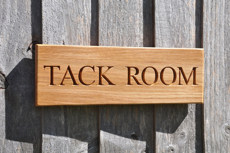 May include: A wooden sign with the words "TACK ROOM" carved into it. The sign is mounted on a gray wooden wall.