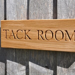 May include: A wooden sign with the words "TACK ROOM" carved into it. The sign is made of light brown wood and has a slightly weathered look.