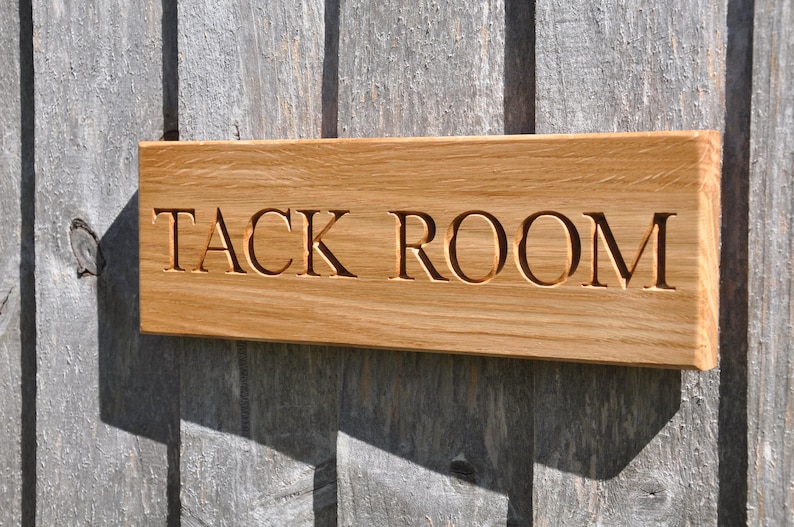 May include: A wooden sign with the words "TACK ROOM" carved into it. The sign is made of light brown wood and has a slightly weathered look.
