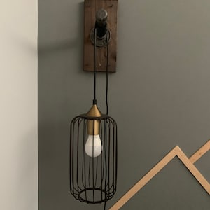 May include: Industrial-style wall sconce featuring a dark metal cage shade with a brass-colored top, housing a white lightbulb. The fixture is mounted on a stained wooden board with black piping and wiring, creating a rustic aesthetic.