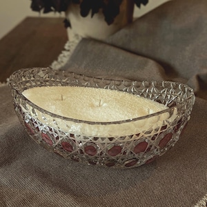 Può includere: Un portacandele grande, di forma ovale, in vetro trasparente con un motivo testurizzato a forma di diamante. Il portacandele contiene una candela bianca ed è posto su una superficie di tessuto marrone.