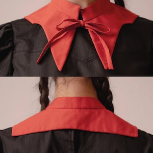 May include: A red detachable collar with a bow detail, worn over a black garment. The collar has a pointed shape and a contrasting colour against the dark fabric. The image shows the front and back of the collar.