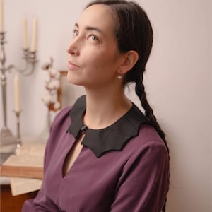 May include: A woman in a plum-coloured dress with a black scalloped collar. The dress has long sleeves and a keyhole neckline. The text "STITCH MAIDEN" is in the upper right corner. The woman has dark hair in plaits and is wearing pearl earrings.