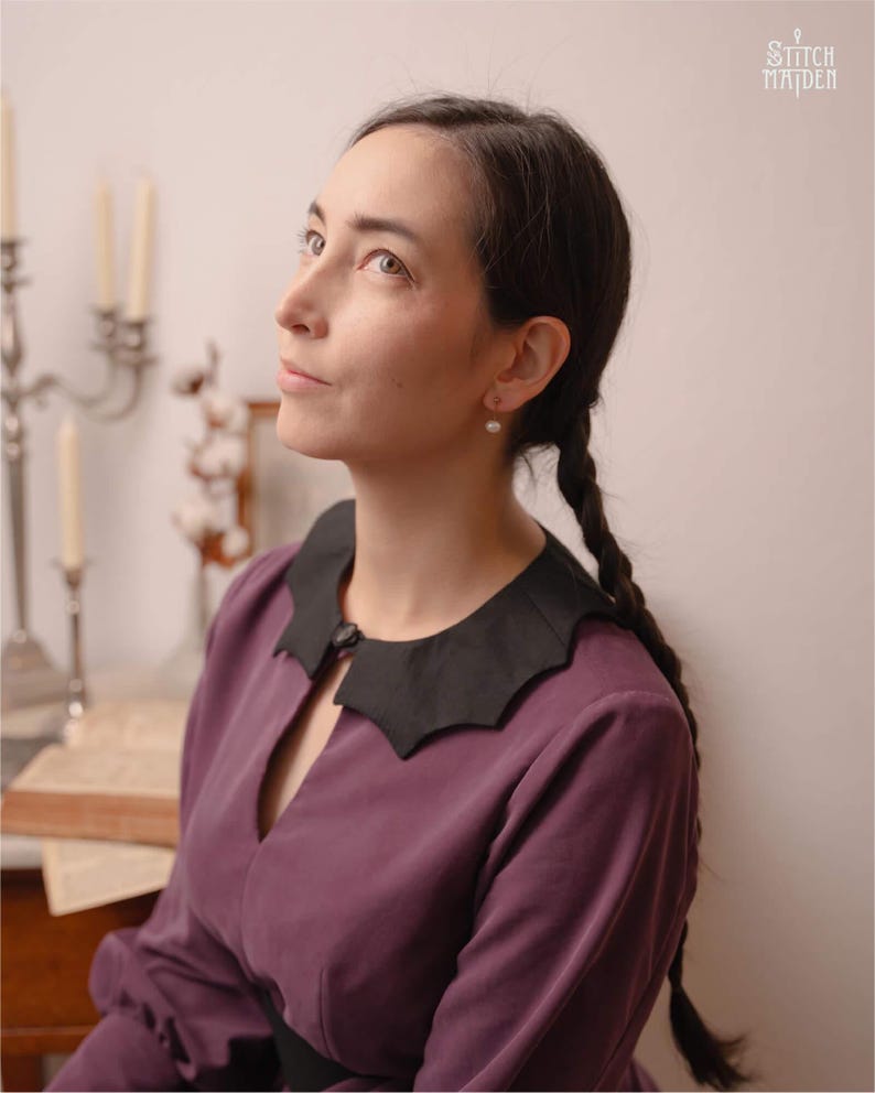 May include: A woman in a plum-coloured dress with a black scalloped collar. The dress has long sleeves and a keyhole neckline. The text "STITCH MAIDEN" is in the upper right corner. The woman has dark hair in plaits and is wearing pearl earrings.