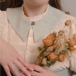 May include: A pale green, textured collar with a decorative button fastening. The collar is worn over a cream blouse with lace detailing. A bouquet of dried flowers is held.