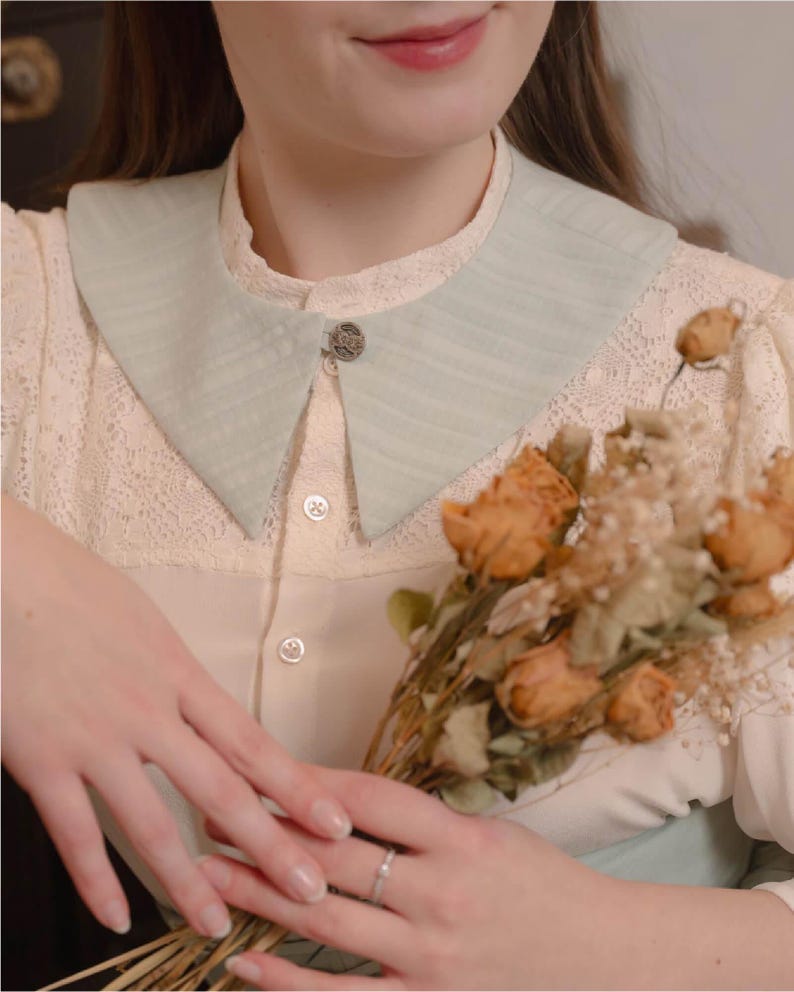 May include: A pale green, textured collar with a decorative button fastening. The collar is worn over a cream blouse with lace detailing. A bouquet of dried flowers is held.